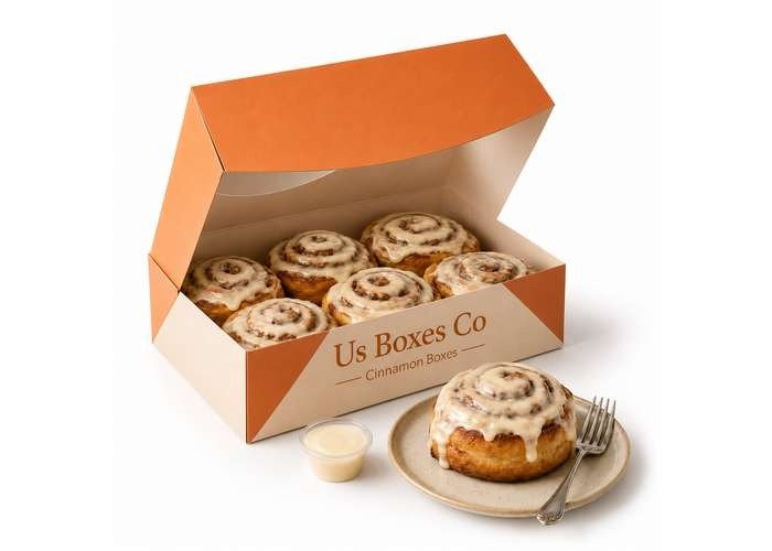 Open orange bakery box filled with iced cinnamon rolls, with one roll served on a plate beside it.